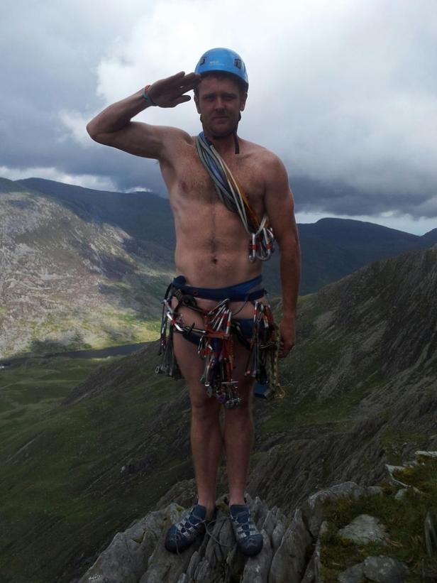 Ein Bergsteiger mit Helm und Kletterausrüstung grüßt auf einem Berggipfel.