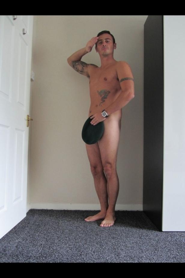 Ein Mann mit Tätowierungen salutiert, während er einen Hut vor seinen Genitalien hält.