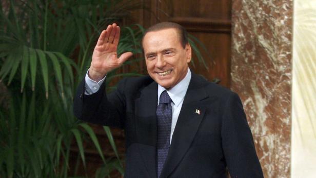 Silvio Berlusconi lächelt und winkt in einem dunklen Anzug.