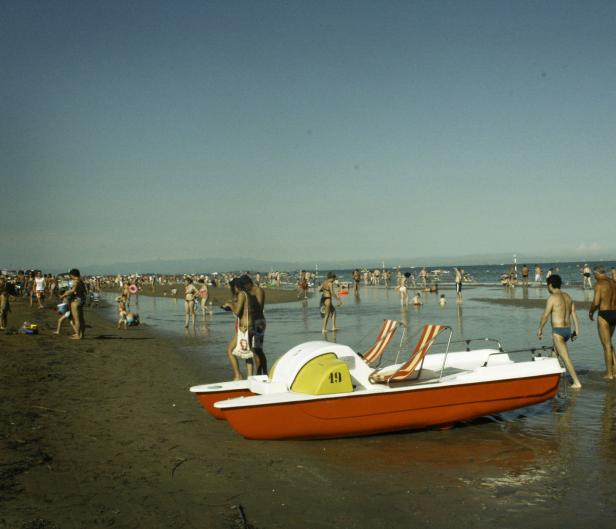 Ein rotes Tretboot liegt am Strand, während sich im Hintergrund viele Menschen im Wasser aufhalten.