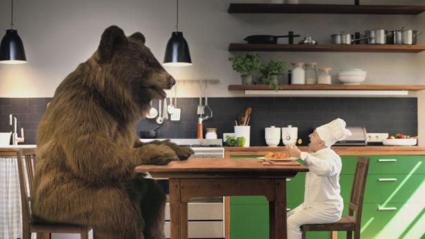 Ein Koch isst mit einem Bären an einem Tisch in einer Küche.