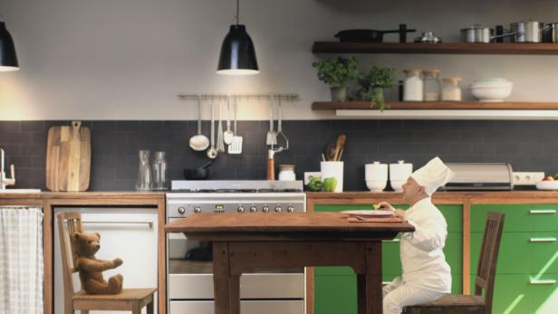 Ein Koch sitzt an einem Holztisch in einer Küche, während ein Teddybär auf einem Stuhl daneben sitzt.