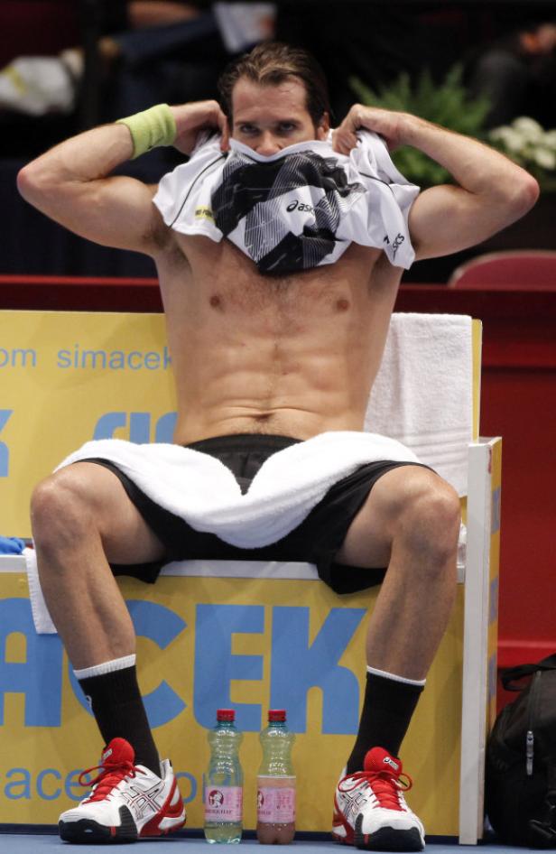 Ein Tennisspieler sitzt während einer Pause auf einer Bank und zieht sein T-Shirt aus.