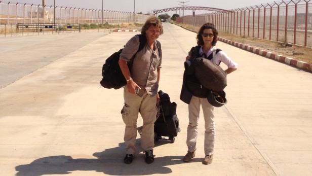 Zwei Reisende stehen mit Gepäck auf einer leeren Straße an einem sonnigen Tag.