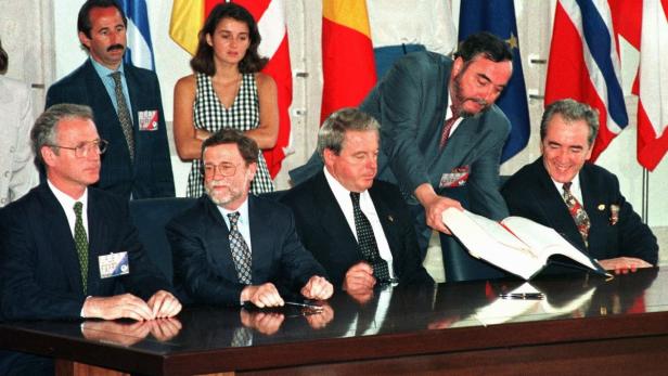 Mehrere Männer in Anzügen sitzen an einem Tisch vor verschiedenen Flaggen.