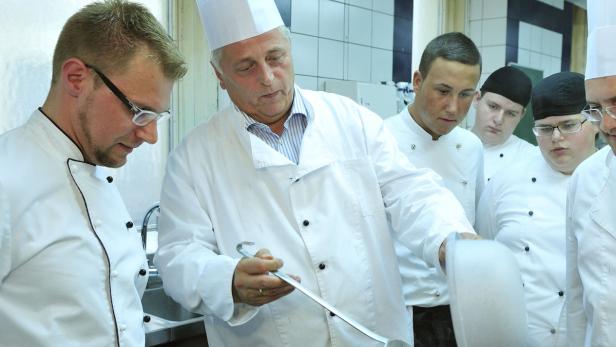 Ein Koch mit Kochlöffel und mehrere Auszubildende in einer Küche.