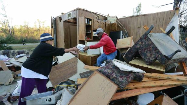 Zwei Personen stehen vor den Trümmern eines Hauses und reichen sich Gegenstände.