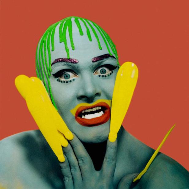 Ein blau geschminkter Mann mit grünen Haaren und gelben Akzenten posiert vor einem roten Hintergrund.