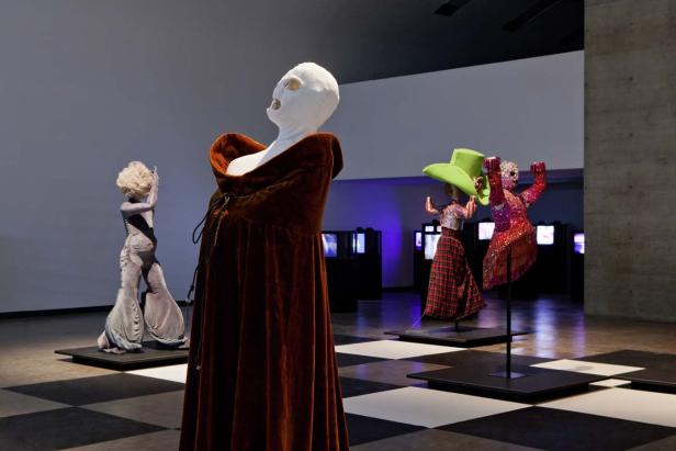 Eine Ausstellung mit skulpturalen Figuren in extravaganten Kostümen auf einem Schachbrettmusterboden.
