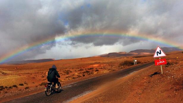 Eine Person fährt mit einem Moped unter einem Regenbogen durch eine Wüstenlandschaft.