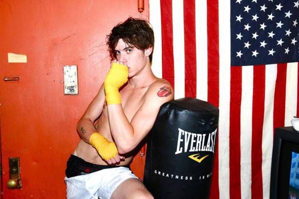 Ein junger Mann mit Boxbandagen posiert vor einem Boxsack und einer US-amerikanischen Flagge.