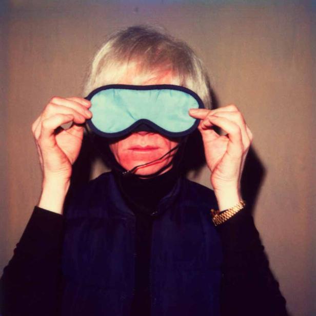 Andy Warhol hält eine hellblaue Schlafmaske vor sein Gesicht.