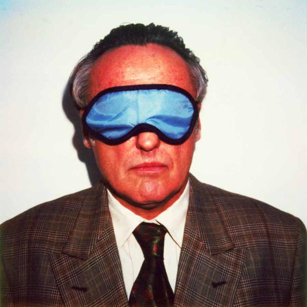 Ein Mann mit Schlafmaske und Anzug blickt in die Kamera.