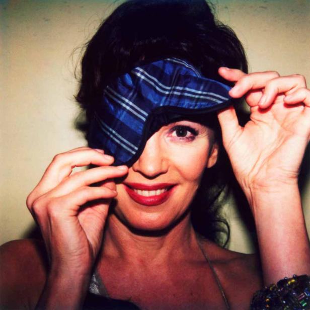 Eine Frau mit rotem Lippenstift hält eine blau karierte Schlafmaske vor ihrem Auge.