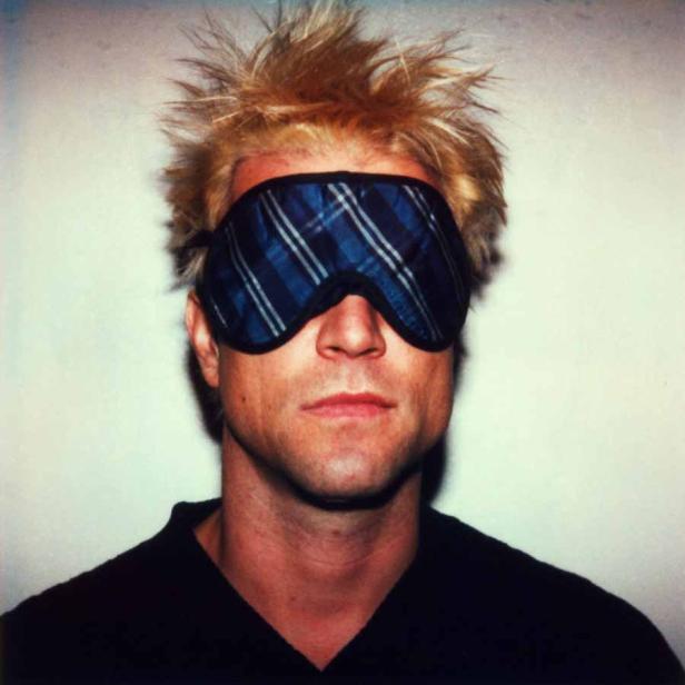 Ein Mann mit blonden, stacheligen Haaren trägt eine blau karierte Schlafmaske.