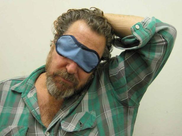 Ein Mann mit Schlafmaske und kariertem Hemd hat eine Hand hinter dem Kopf.