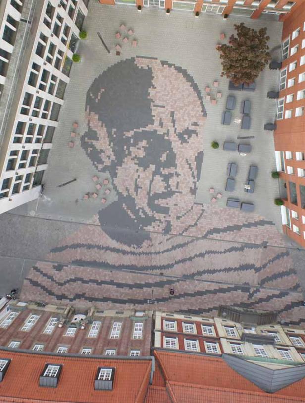 Ein aus Pflastersteinen gestaltetes Porträt auf einem Platz zwischen Gebäuden, aufgenommen aus der Vogelperspektive.