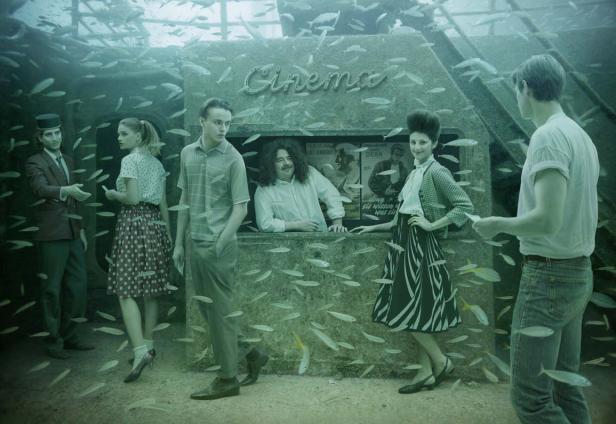 Eine Gruppe von Menschen steht vor einem versunkenen Kino, umgeben von Fischen.