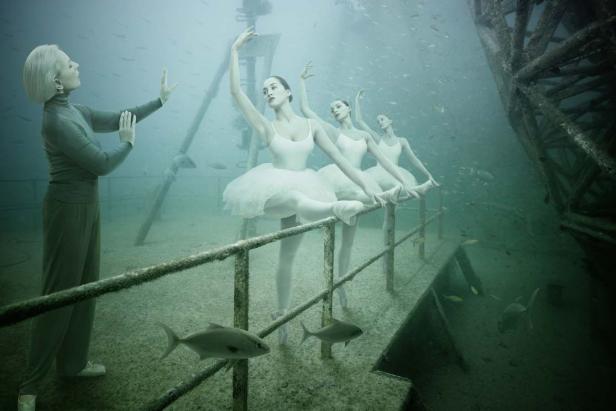 Eine Ballettlehrerin dirigiert drei Ballerinen unter Wasser vor einem versunkenen Schiffswrack.