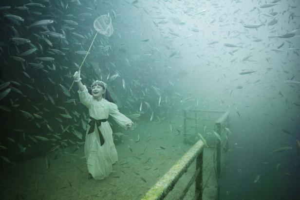 Ein Mädchen mit einem Fischernetz steht inmitten eines Schwarms Fische unter Wasser.