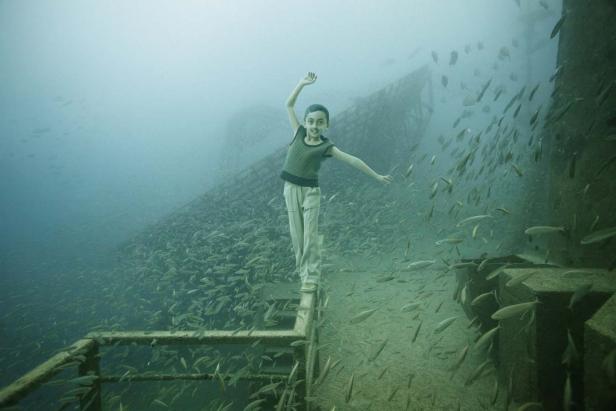 Ein Kind steht auf einer Treppe in einem versunkenen Schiff, umgeben von einem Fischschwarm.