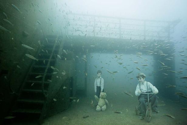 Zwei Jungen in altmodischer Kleidung und ein Teddybär in einer Unterwasserumgebung mit vielen Fischen.