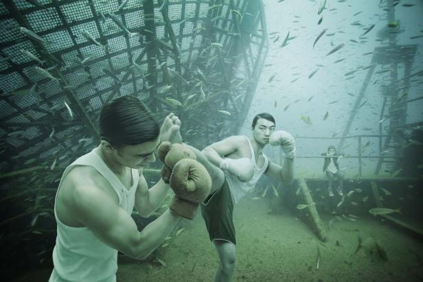 Zwei Männer boxen unter Wasser, umgeben von Fischen und einer versunkenen Struktur.