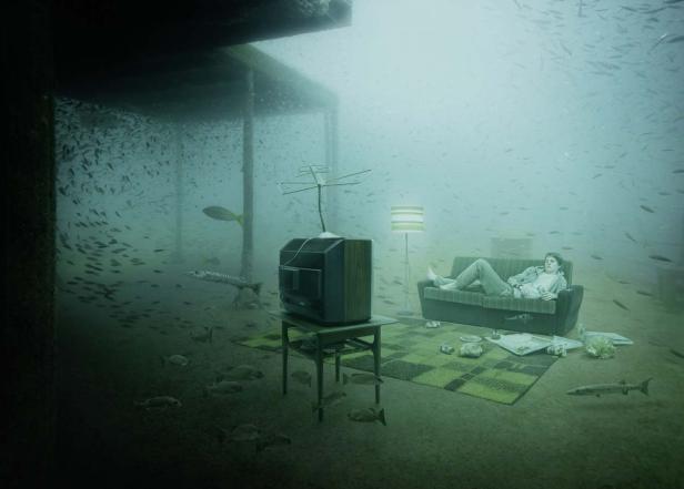Ein Mann liegt auf einem Sofa unter Wasser, umgeben von Fischen und einem Fernseher.