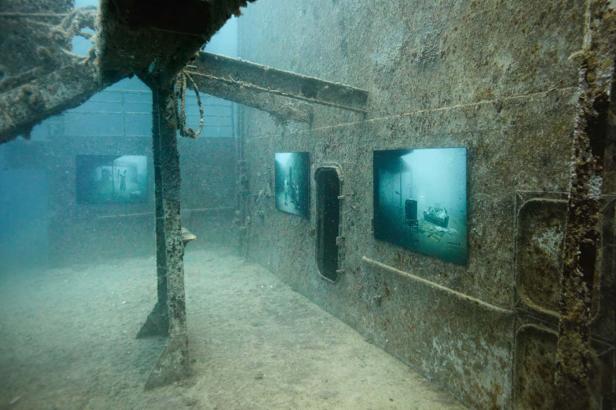 Unterwasseraufnahme eines Schiffswracks mit angebrachten Fotografien an den Wänden.