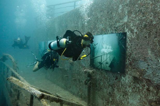 Zwei Taucher betrachten ein Unterwasser-Kunstwerk an der Wand eines Schiffswracks.