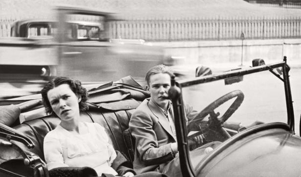 Ein Mann und eine Frau sitzen in einem offenen Oldtimer, während ein anderes Auto vorbeifährt.