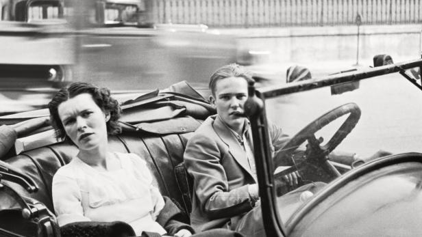 Ein Mann und eine Frau sitzen in einem Oldtimer, während im Hintergrund der Verkehr vorbeifließt.