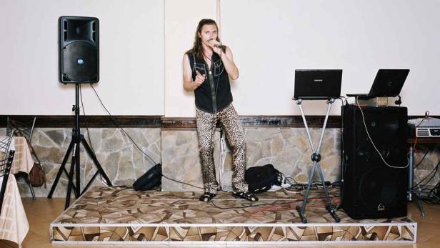 Ein Mann mit Leopardenhose singt auf einer Bühne mit Lautsprechern und Laptops.