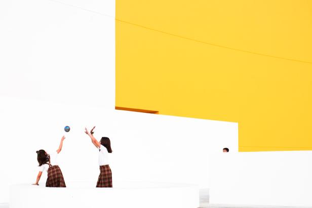 Zwei Mädchen in Schuluniform spielen mit einem Ball vor einer gelb-weissen Wand.