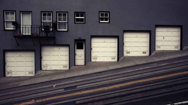 Eine steile Straße führt an einer grauen Hausfassade mit Garagentoren und Fenstern vorbei.