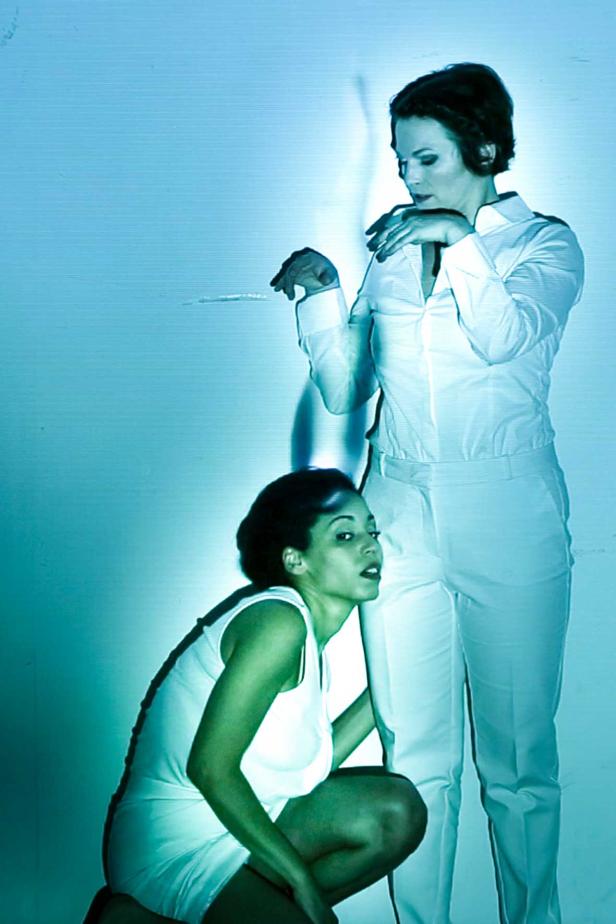 Eine stehende Frau im weißen Anzug dirigiert eine kauernde Frau mit einem Taktstock.