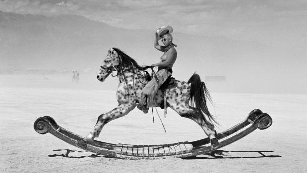 Eine Frau im Cowboy-Outfit reitet auf einem Schaukelpferd in einer Wüstenlandschaft.