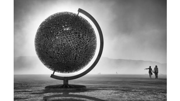 Eine große Globus-Skulptur steht in einer nebligen Wüstenlandschaft, im Hintergrund ein Paar.