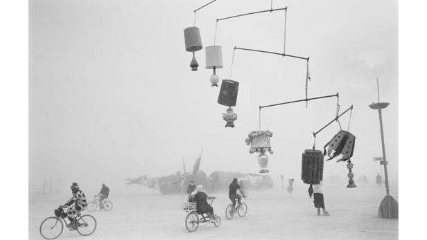 In einer staubigen Landschaft fahren Menschen mit Fahrrädern unter einem Mobile aus Lampenschirmen hindurch.
