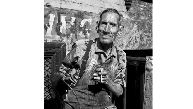 Ein älterer Mann mit Hosenträgern und Schürze hält eine Zigarette in der Hand.