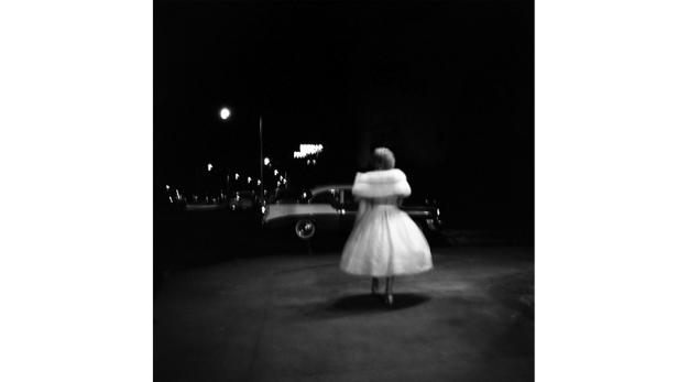 Eine Frau in einem Kleid geht nachts an einem Oldtimer vorbei.