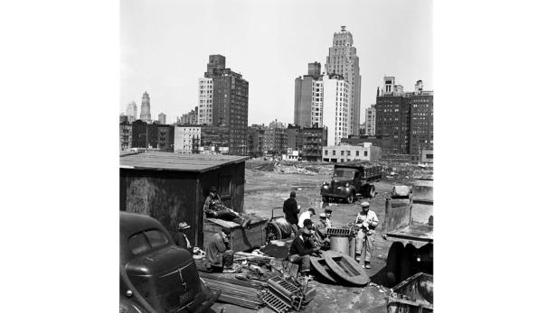 Eine Gruppe von Männern pausiert auf einer Baustelle in einer Stadtlandschaft.