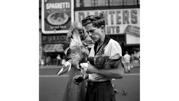 Ein junger Mann füttert Tauben auf einer belebten Straße in Schwarzweiß.