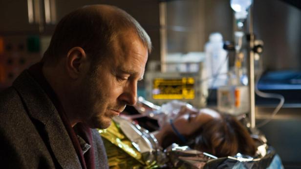 Ein besorgter Mann blickt auf eine bewusstlose Frau, die an medizinische Geräte angeschlossen ist.