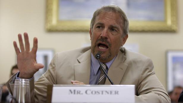 Kevin Costner spricht bei einer Anhörung und hebt seine Hand.