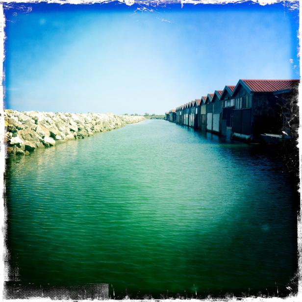Ein Kanal mit türkisfarbenem Wasser, begrenzt von einem Steindamm und einer Reihe von Bootshäusern.
