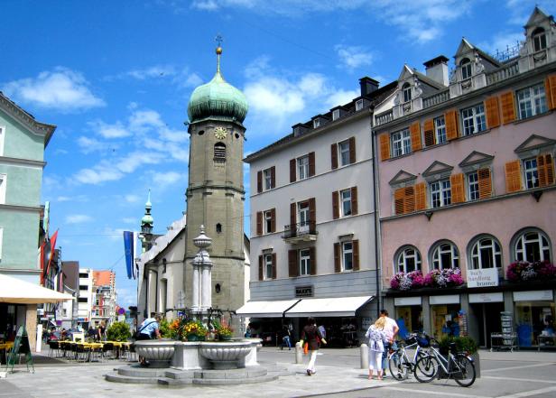 Ein belebter Platz in einer europäischen Stadt mit einem Brunnen und einem Kirchturm.