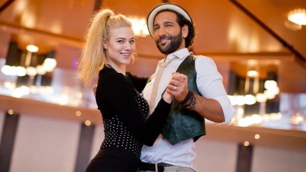 Ein Mann mit Hut und eine blonde Frau tanzen zusammen in einem Ballsaal.