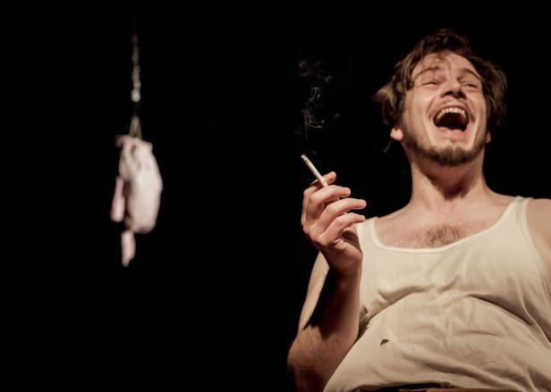 Ein lachender Mann mit Unterhemd raucht eine Zigarette; im Hintergrund hängt ein Stück Fleisch.