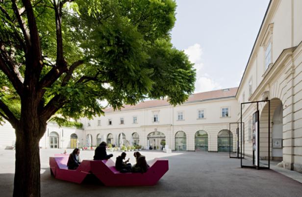 Menschen sitzen auf einer pinkfarbenen Bank im Hof eines Gebäudes.